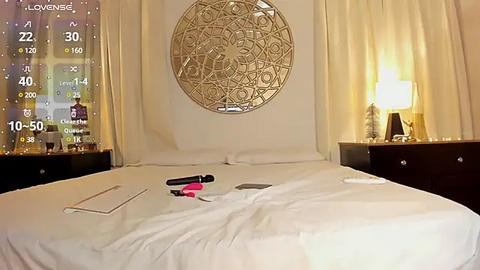 Media: Video of a luxurious, modern hotel room with a circular, metallic wall art piece above the bed, cream-colored curtains, and a white bed with a black iPhone, black and pink lipstick, and a tablet on top.