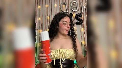 Media: Video of a young woman with curly dark hair, wearing glasses and a yellow strapless top, holding a red cup. Background features silver confetti, \"18\" letters, and festive lights.