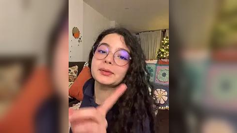 Media: Video of a young woman with curly black hair and large round glasses, making a peace sign, in a cozy living room with a Christmas tree and colorful throw pillows.