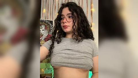 Media: A video of a young woman with curly dark hair, wearing glasses, a grey crop top, and standing against a backdrop of colorful posters and fairy lights.