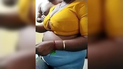 Media: Video of a pregnant woman in a tight, mustard-yellow crop top and blue pants, holding her baby, blurred background.