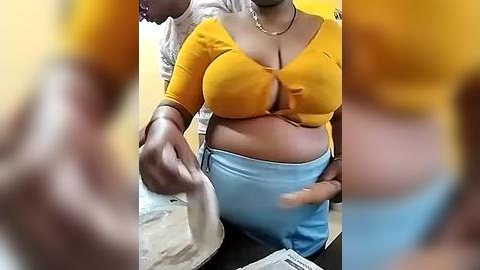 Media: Video of a dark-skinned woman with a large bust, wearing a yellow crop top and light blue pants, holding a white plastic bag, blurred background.