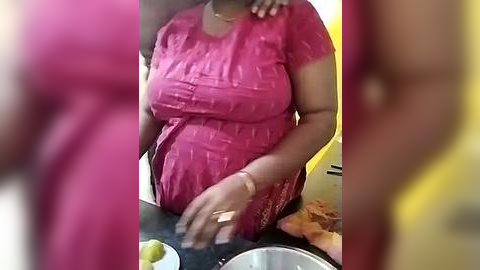 Media: Video of a woman with medium-dark skin, wearing a pink, short-sleeved, textured dress, standing in a kitchen. She is holding a green apple. Background includes a yellow wall and a black countertop.