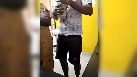 Media: A video of a person with dark skin and a prosthetic leg, wearing a white patterned shirt and black shorts, taking a selfie in a brightly lit, yellow-walled bathroom with a toilet and sink in the background.