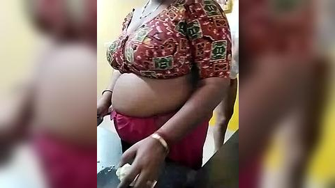 Media: Video of a pregnant woman with medium brown skin, wearing a red and green patterned crop top and red pants, standing indoors.