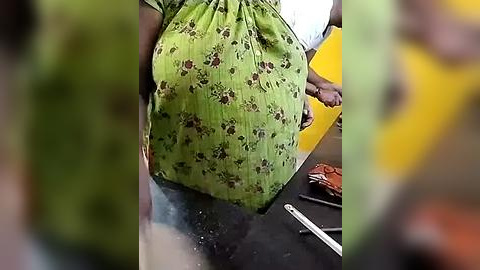 Media: Video of a pregnant woman wearing a green dress with brown floral patterns, seated on a dark bench. Her belly is prominent. Background includes blurred figures and objects.