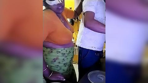 Media: Video of two women in a kitchen; one wearing an orange shirt, green patterned skirt, and a mask, the other in a white top and blue jeans.