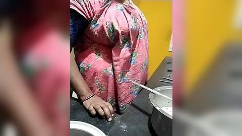 Media: Video of a woman with a medium complexion, wearing a pink floral dress, bending over a sink, hands in water. Background includes a yellow wall and a blurred figure.