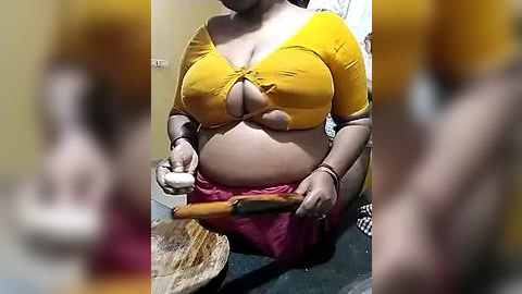 Media: Video of a pregnant woman in a yellow crop top, red skirt, holding a wooden rolling pin in a kitchen. Background blurred.