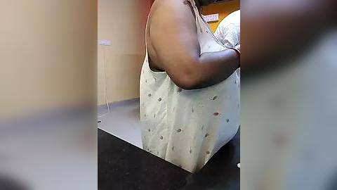 Media: A video of a dark-skinned person with a muscular build, wearing a sleeveless white shirt, carrying a light-colored tote bag with small, dark patterns. The background shows a beige wall and a partially visible bed.