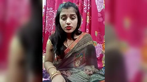 Media: Video of a South Asian woman with medium skin tone and long black hair, wearing a traditional sari with intricate patterns, sitting in a brightly colored room with red curtains and floral patterns.