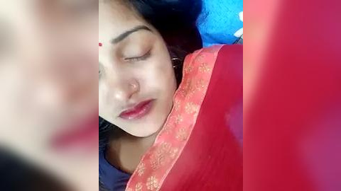 Media: Video of a serene South Asian woman with fair skin, closed eyes, and red dot on forehead, wearing a red sari with golden embroidery, lying on a blue surface.