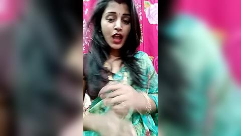 Media: Video of a young woman with long black hair, wearing a turquoise saree, seated in a pink room, looking distressed, blurred background.