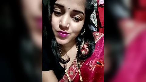 Media: Video of a young woman with medium skin tone, wearing a red sari with gold embroidery, smiling, with a nose ring, and wearing pink lipstick.