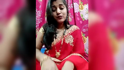 Media: Video of a woman in a vibrant red sari with intricate gold embroidery, sitting against a backdrop of pink floral curtains. She has long black hair and a serene expression.