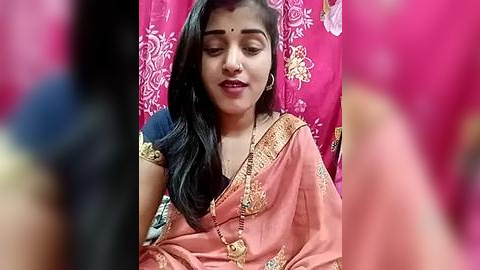 Media: Video of a young Indian woman with medium skin tone, wearing an ornate pink saree with gold embroidery, sitting in a chair against a pink background with white floral patterns.