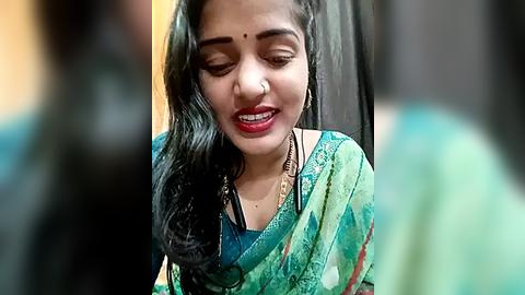 Media: A video of a South Asian woman with long, wavy black hair, wearing a green sari with intricate designs, smiling with red lipstick, against a blurred background.