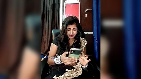 Media: Video of a South Asian woman in traditional attire, black sari with gold embroidery, sitting in a train, engrossed in her smartphone. Background includes dark wooden paneling and a red compartment door.