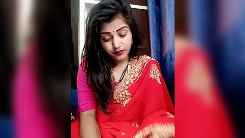 Media: Video of a young South Asian woman with medium skin tone, long black hair, and a red saree with gold embroidery, seated in a dimly lit room with blue curtains.