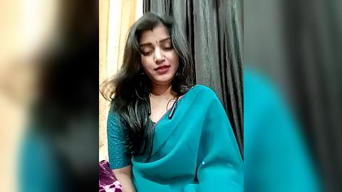 Media: A video of a South Asian woman with long black hair, wearing a teal saree and a purple blouse, standing in front of dark curtains.