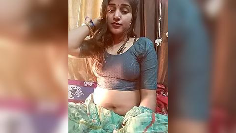 Media: A video of a South Asian woman with medium brown skin and long dark hair, wearing a blue crop top and green sari, sitting on a bed with a blurred background.