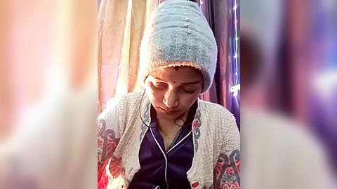 Media: Video of a young woman with light skin, wearing a light blue knit hat, purple headband, and a patterned white sweater over a dark top. She has closed eyes and appears contemplative, seated in a dimly lit, blurry background.