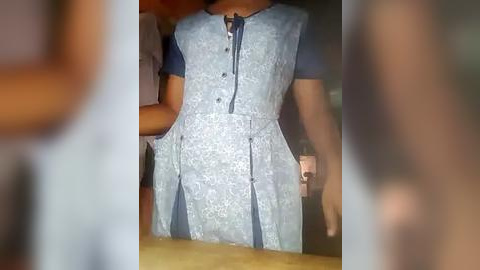 Media: Video of a woman wearing a light blue, lace dress with a button-up front and a small, visible cleavage. Her arms are outstretched, and the background is blurry, showing a wooden surface and part of another person.