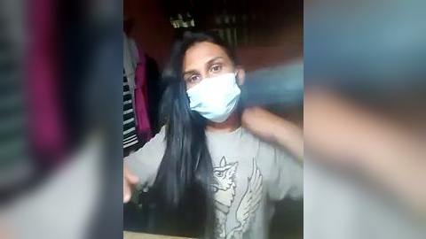 Media: Video of a young woman with long black hair, wearing a light gray t-shirt with a cat design and a blue surgical mask, standing in a dimly lit room with blurred people and objects in the background.