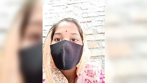 Media: Video of a young woman with a light brown complexion, wearing a black face mask, light floral blouse, and a white headscarf against a brick wall.