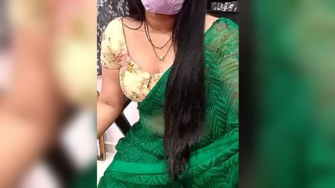 Media: Video of a woman with long black hair, wearing a lavender mask, green sari with floral blouse, sitting on a chair with a blurred background.