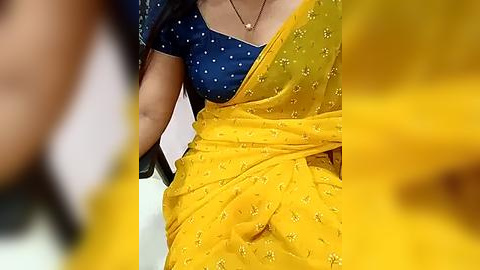 Media: Video of a woman wearing a blue blouse with white polka dots and a vibrant yellow saree with gold embroidery, hands resting on a black chair.