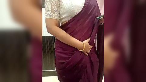 Media: A video of a woman in a maroon sari with a white lace blouse, holding the sari's edge with her right hand. She wears a gold bracelet. Background is blurred, showing a partially visible person in a dark outfit.