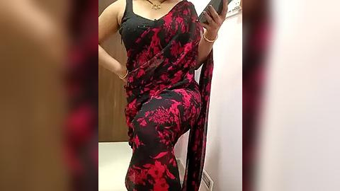 Media: A video of a woman in a black and red floral sari with a black sleeveless blouse, standing in a bathroom with beige tiles and a wooden door background.