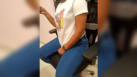 Media: Video of a seated person with medium brown skin, wearing a white graphic t-shirt and blue jeans, occupying a black office chair in a minimalistic room.