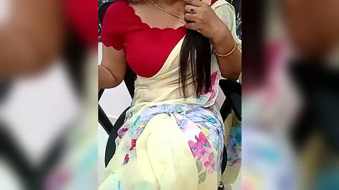 Media: Video of a woman with medium brown skin, wearing a red blouse and a pastel floral saree, sitting in a chair, her long, straight hair partially covering her face.