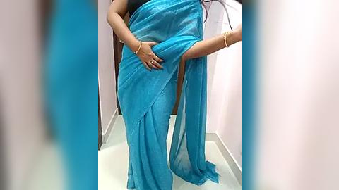 Media: A video showing a person in a flowing, light blue saree, with their arm wrapped around their torso, standing in a pastel-painted room with a white floor.