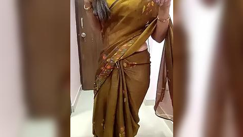 Media: Video of a woman in a gold sari with floral patterns, holding a pink cloth, standing in a corridor with light walls and a door.