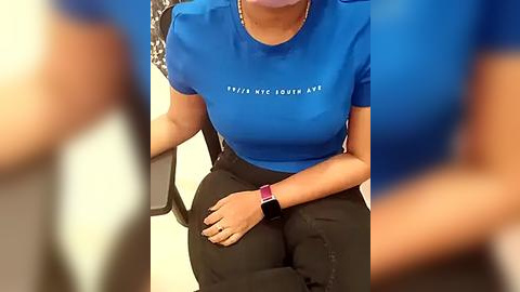 Media: Video of a woman in a bright blue t-shirt with white text, black pants, and a red watch, seated in a chair.