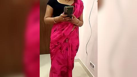 Media: A video of a woman in a bright pink saree with black blouse, holding a smartphone, standing in a narrow hallway with a white wall and beige floor.