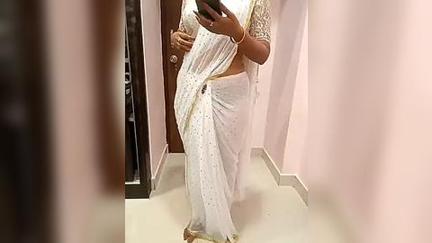 Media: Video of a person wearing a white, embroidered sari, with intricate gold patterns, and a matching blouse, standing in a pink-walled, modern room with a tiled floor.