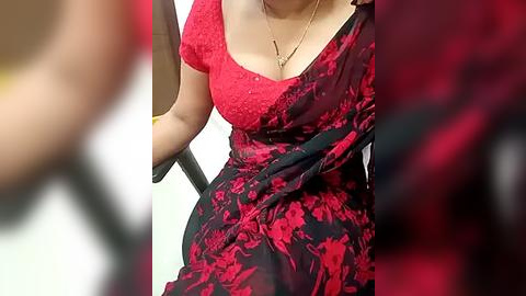 Media: Video of a woman wearing a red and black floral sari with a gold necklace, sitting in a chair. The background is blurred.