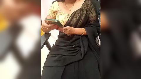 Media: Video of a woman in a black saree with a gold blouse, holding a smartphone, blurred background, possibly indoors.