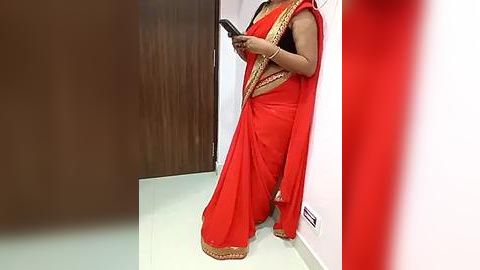 Media: Video of a woman in a bright red saree with gold trim, holding a smartphone, standing against a white wall with a brown door and tiled floor.