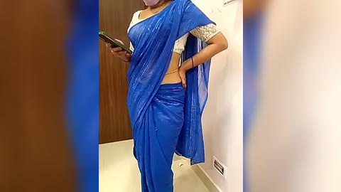 Media: Video of a woman in a vibrant blue sari with white lace trim, holding a smartphone, standing in a hallway with wooden door and white walls.