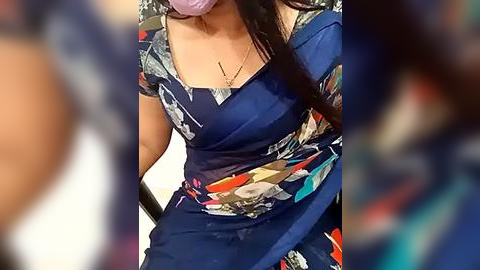 Media: Video of a woman with long dark hair, wearing a blue floral dress with a plunging neckline, seated indoors. The background is blurred, highlighting her attire and necklace.