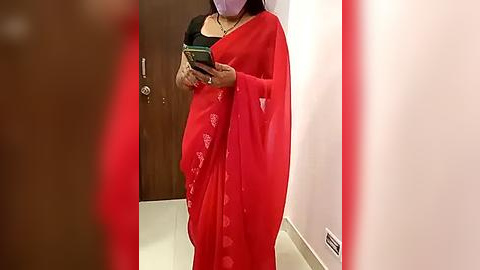 Media: A video of a pregnant woman in a bright red saree with white floral patterns, holding a smartphone, standing in a hallway with light beige tiles and a dark wooden door.