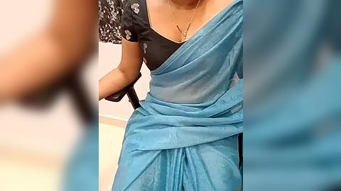 Media: Video of a woman in a black blouse with white floral embroidery, wearing a light blue saree with a gold necklace, walking with a cane.