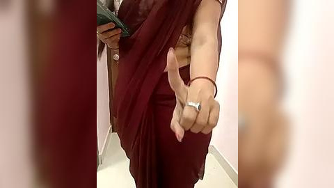 Media: A video captures a woman in a maroon sari, holding a green bag, and making a peace sign with her right hand, wearing a silver ring. The background is a plain, light-colored wall.