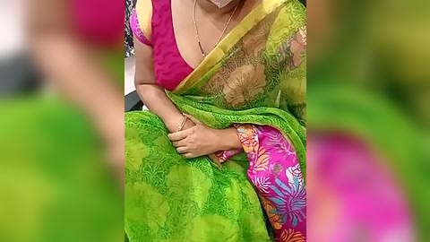 Media: Video of a woman in a bright green sari with pink floral patterns, seated, wearing a pink blouse, and holding a phone.