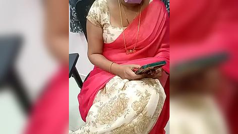 Media: Video of a woman in a bright red sari with intricate golden patterns, seated on a black chair, holding a smartphone, wearing a gold necklace, in a modern indoor setting.